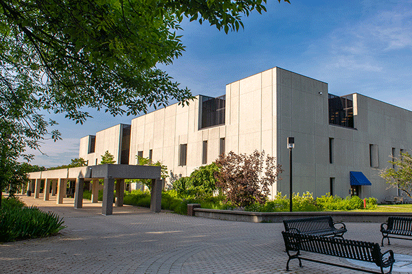 white, multi-story square building in the distance with meal benches surrounding an ISU seal on the ground and a walkway leading to the building. A tree with green leaves is on the right with blue sky above.